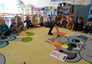 Dziewczynka układa pajaca z figur geometrycznych według wzoru, dzieci siedzą wokół niego i obserwują z zainteresowaniem.